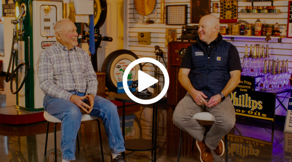 image of two men sitting on stools talking with a play button over the top